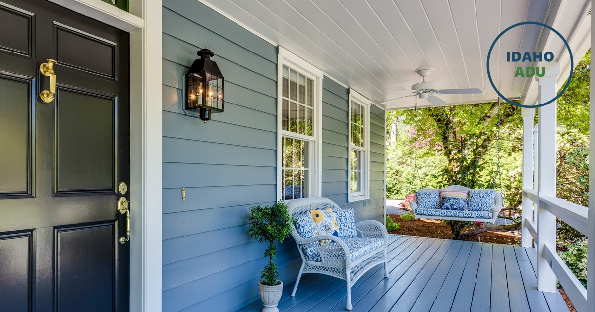 The covered front porch of a blue clapboard house, dark front door with brass hardware, a white wicker chair and a porch swing