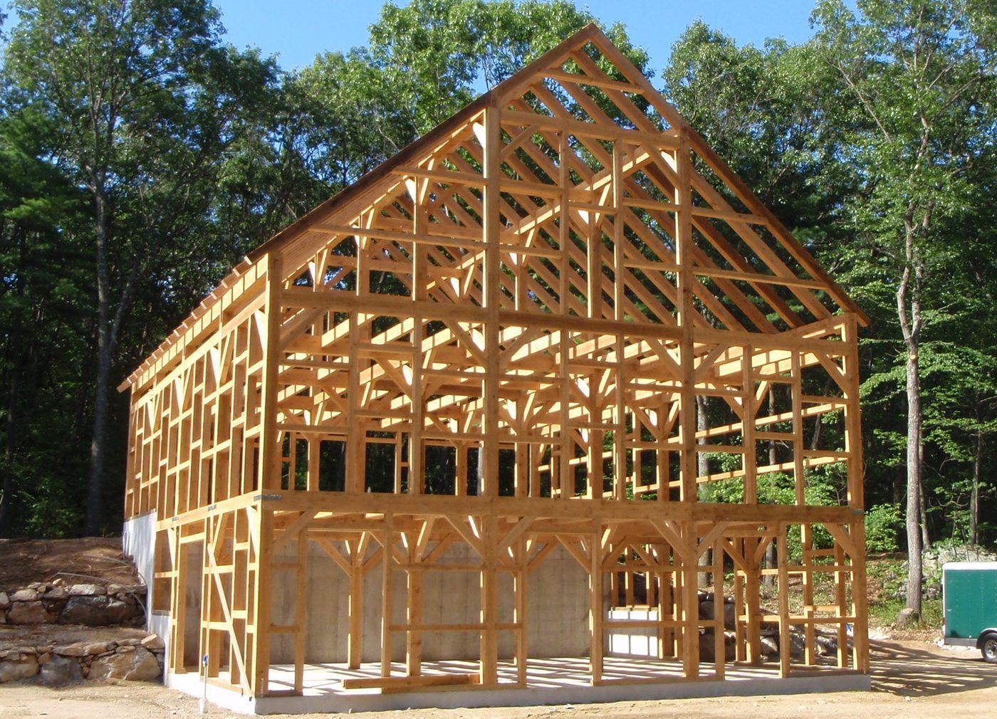 A timber frame skeleton just raised in a clearing: honey-colored posts, beams and diagonal braces, roof rafters up, no walls yet, trees behind it.